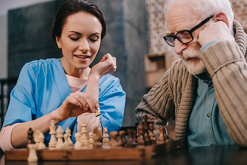 Young female nurse in blue scrubs playing chess with older gentleman wearing glasses