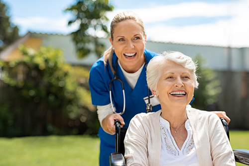 CNA in blue scrubs pushing elderly smiling woman in wheelchair outside